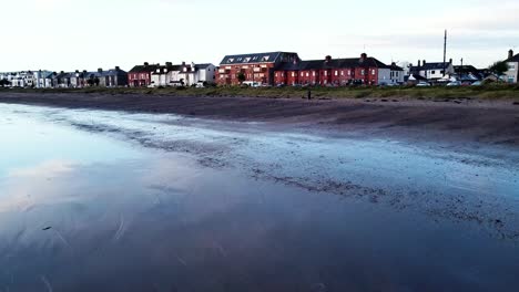 A-low-drone-shot-passing-over-the-sandy-coastal-shore-of-Skerries-beach-during-low-tide-approaching-the-coastal-residential-houses,-Ireland