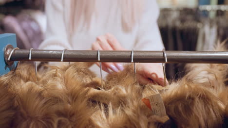 hand view of woman shifting winter coat on hanger sliding it aside on rack while feeling fur trim and inspecting fabric quality with manicured nails under warm boutique lighting