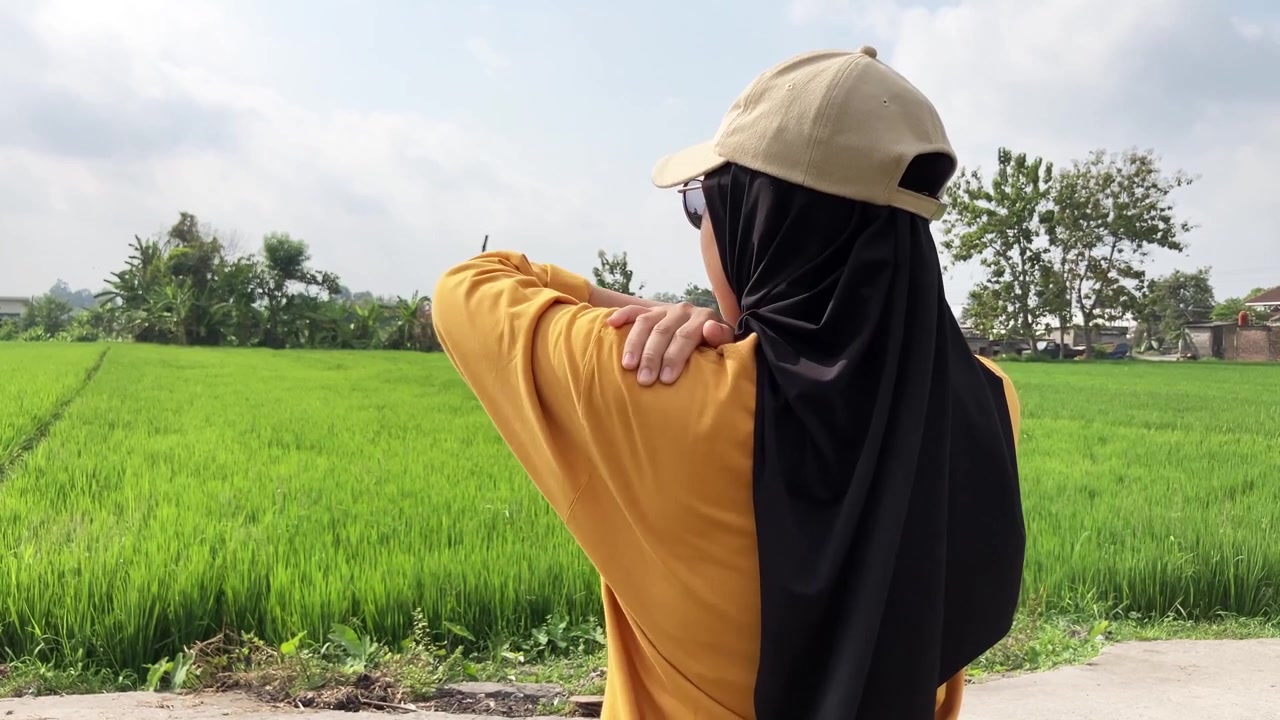 Tampilan belakang gerak lambat seorang wanita Muslim berhijab dan bertopi memegangi bahu kirinya kesakitan. Dia mengalami nyeri otot saat berdiri di luar di samping sawah hijau subur