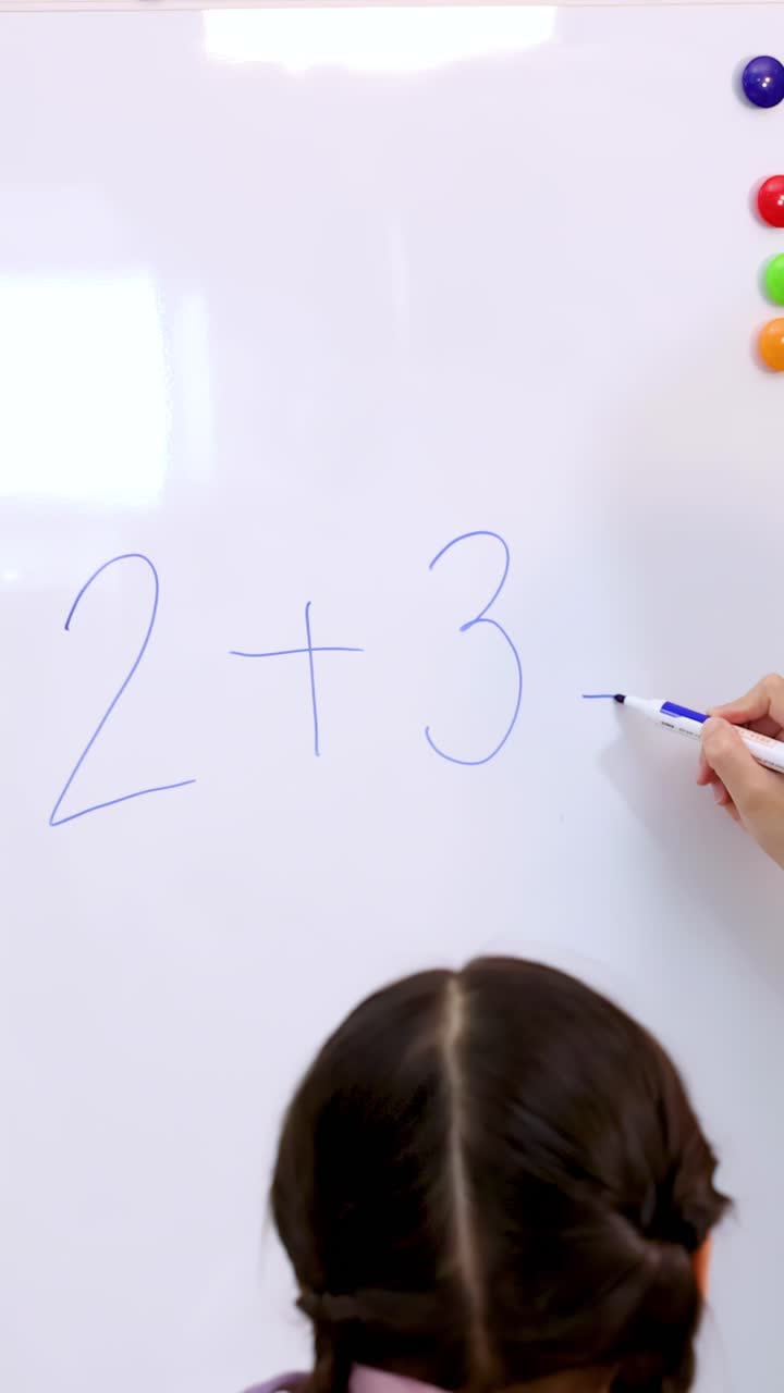 A teacher writes an addition problem on a whiteboard while a young student watches closely