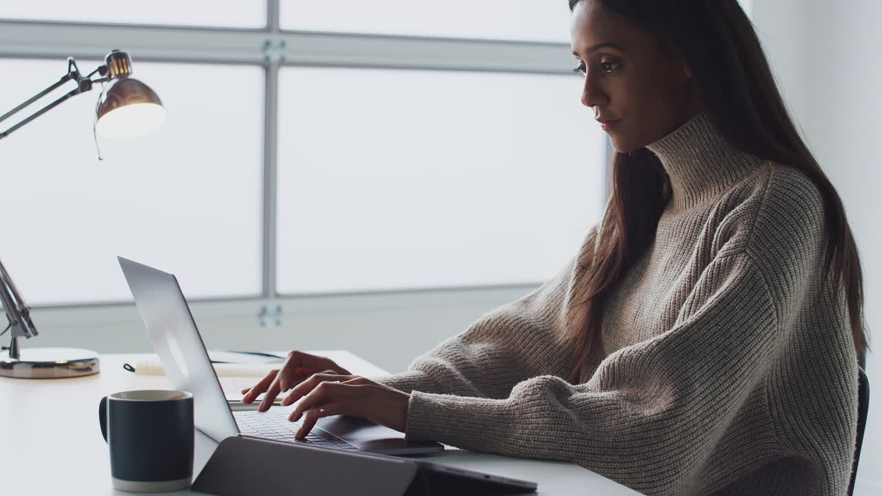 Businesswoman Working On Laptop At Desk In Modern Office Looking At Notebook