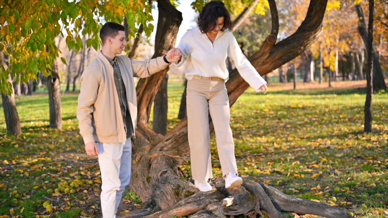 A romantic couple in an autumn park. Man holding his woman while she is walking along a tree trunk. Autumn atmosphere, yellowing trees and leaves around. Slow motion