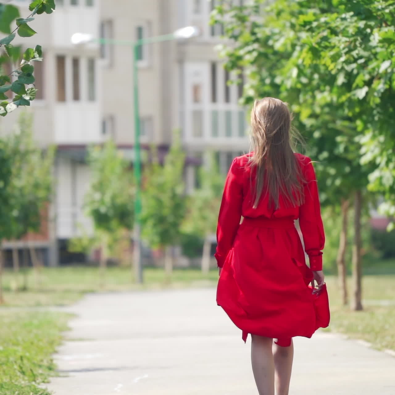 Back view of a woman in summer. Young female in red dress with long hair is walking on the street to the building in the city.Slow motion.