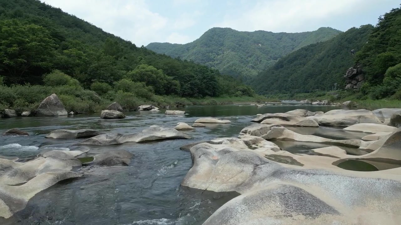 韓国の、豊かな緑の森と山々に囲まれた、滑らかな花崗岩の岩層の上を流れる手つかずの山間の川の広大な風景ショット。透き通った水、壮大な自然の