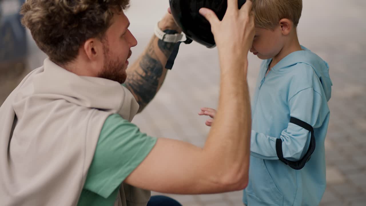 초록색 티셔츠를 입은 곱슬머리 아빠가 여름에 스케이트 파크에서 스케이트보드를 타는 동안 보호를 위해 검은 헬을 착용하는 작은 아들을 도와줍니다.