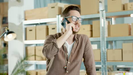 uomo stilista cucasiano in occhiali che parla al telefono e si appoggia al tavolo in un negozio di abbigliamento di moda