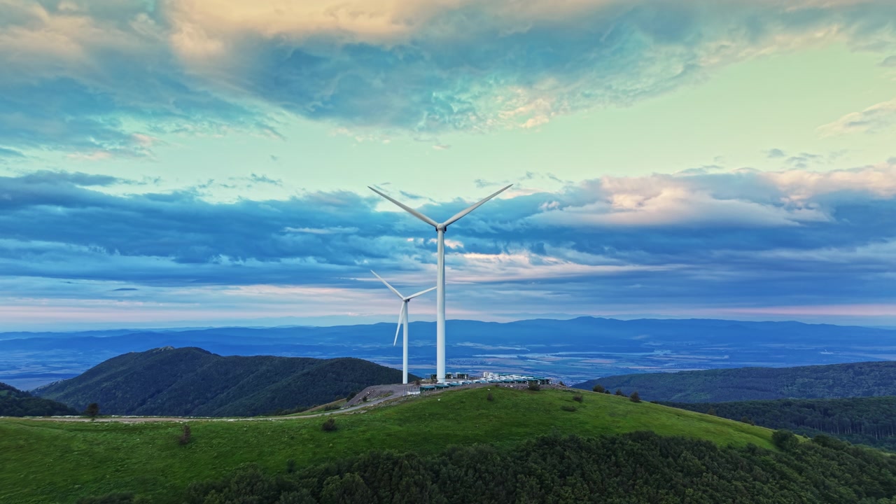 Turbine eoliche si ergono su una collina mentre le nuvole si addensano nel cielo e il sole tramonta in lontananza su un paesaggio verde
