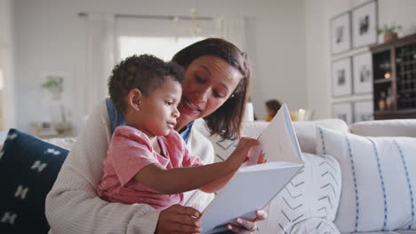 madre afroamericana sentada en el sofá en la sala de estar leyendo a su hijo pequeño, de rodillas, de cerca