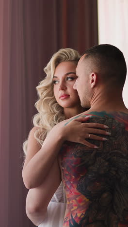 boyfriend with complicated tattoos on back hugs pretty blonde woman standing by large window with curtains in hotel room at honeymoon slow motion