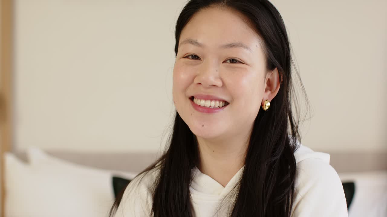 Asian woman in hoodie raising hands, smoothing hair on bed, checking gold earrings and laughing