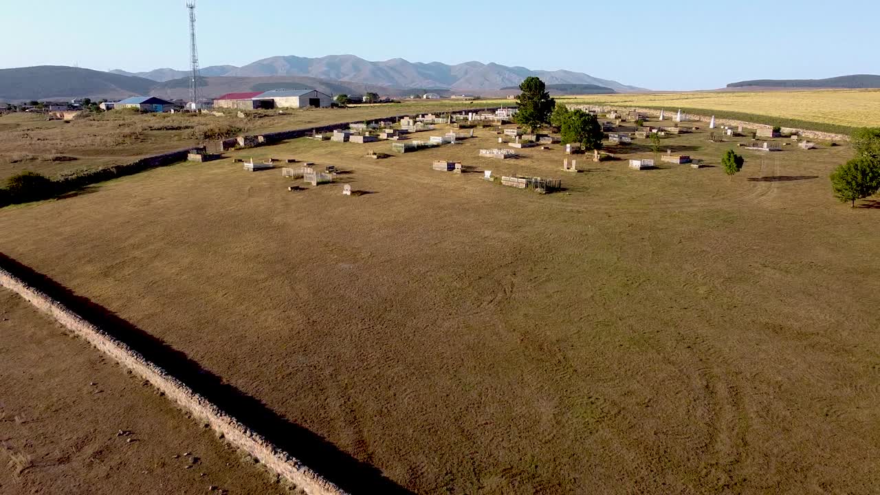 A large abandoned graveyard is located in a peaceful rural area, with few trees and distant mountains. Grave markers are scattered throughout the grassy land, showcasing a sense of history