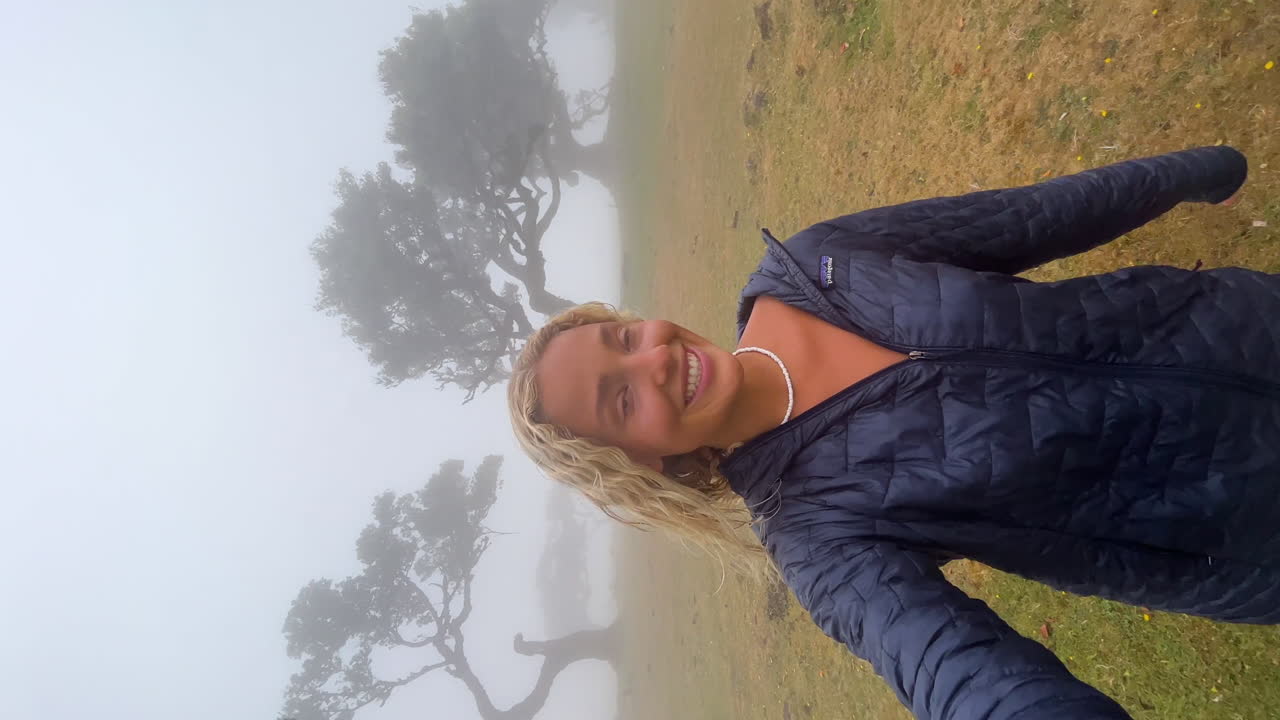 Beautiful blonde girl with a smile on her face filming herself while walking lost in a mystical misty forest