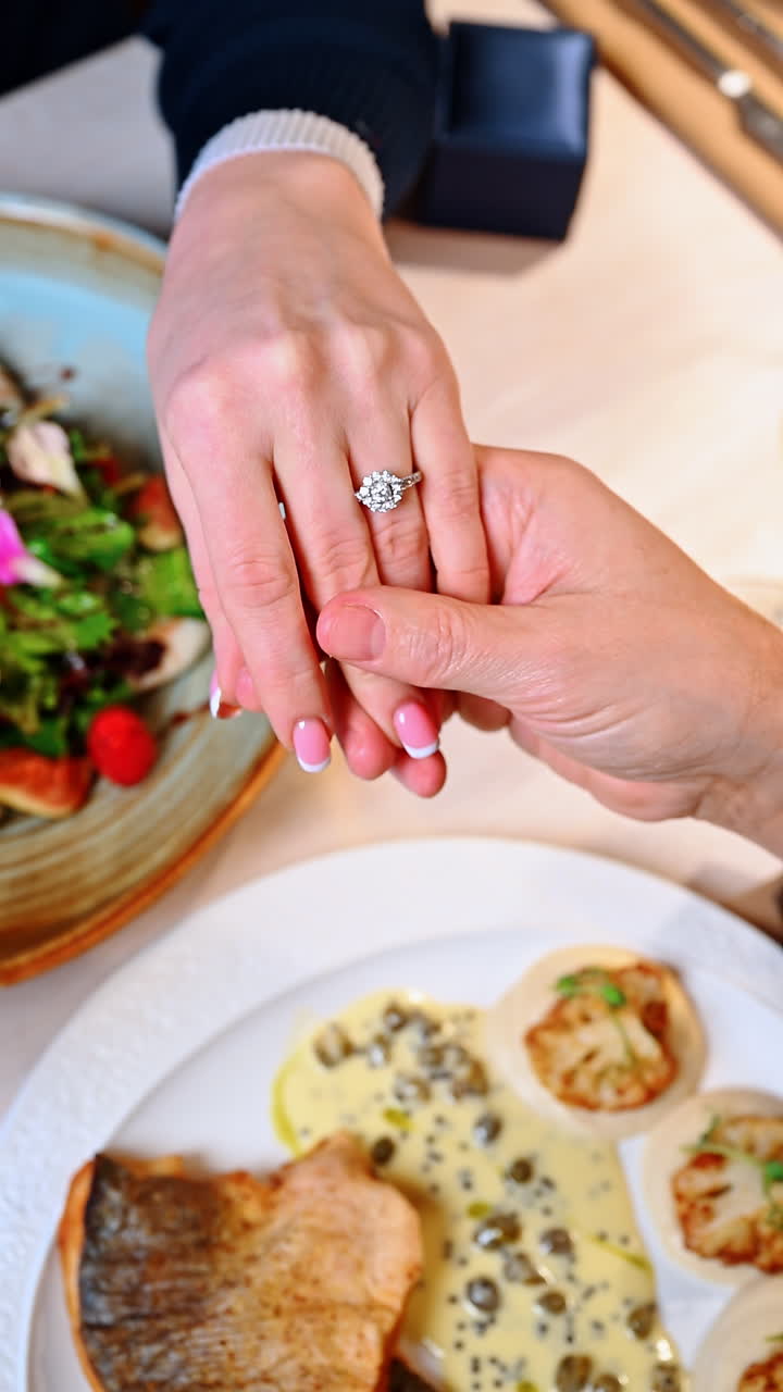 Couple shares a special moment during dinner celebration in a restaurant. Two hands come together over a dinner table with food and drinks, showing a ring on one hand