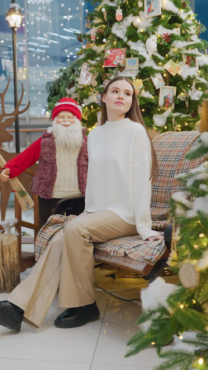 Young woman in brown trousers and white top sits on chair decorated with Christmas decor, surrounded by festive trees and Santa figure, inside a shopping mall decorated for the holiday season
