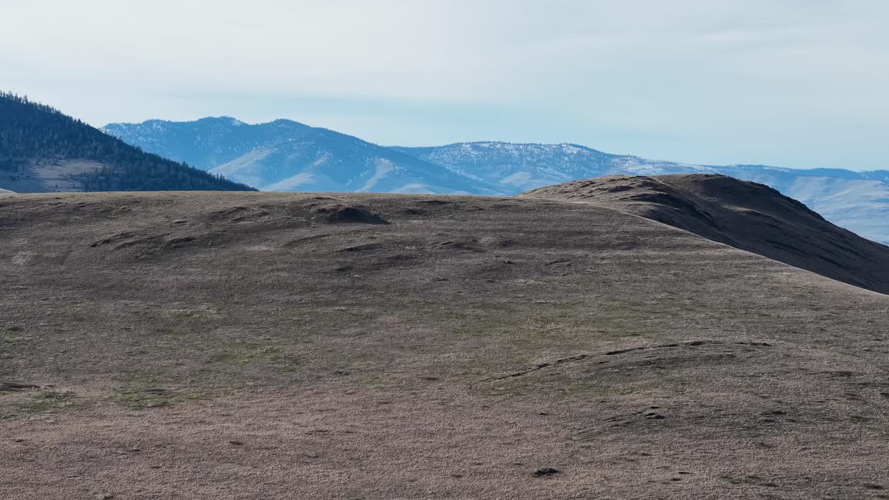 Riprese di drone mostrano un'ampia vista di colline ondulate e aride e una catena montuosa distante sotto un cielo azzurro pallido