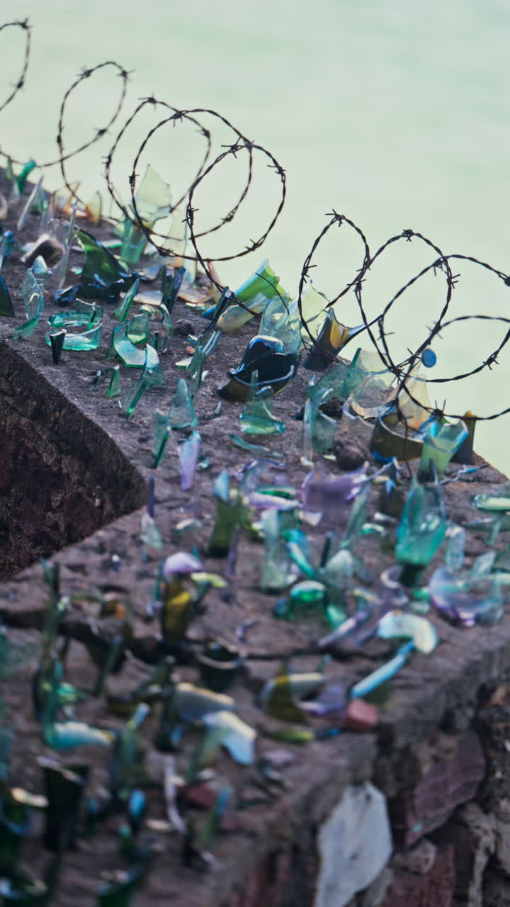 Close up of multiple broken glass shards and barbed wire loops along the edge of a brick wall with a blurred view of the sea. Vertical