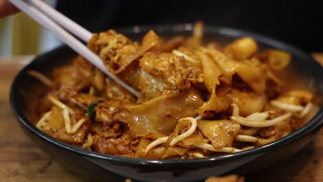 chopsticks lift saucy flat rice noodles, egg, and bean sprouts from a black bowl under warm indoor lighting, highlighting singaporean street food in close-up