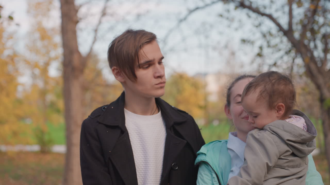 Giovane famiglia caucasica in un parco alberato, la madre tiene stretto il bambino mentre il padre osserva con un sorriso dolce, alberi autunnali sullo sfondo, scatto di vita spontaneo, legame di sostegno, abbigliamento casual, accogliente.
