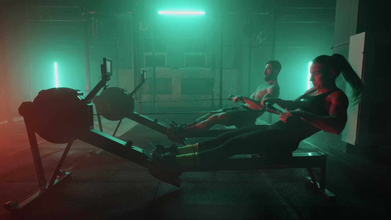 Slow motion: Man and woman training on a rowing machine in gym