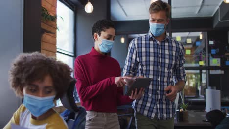 Caucasian-male-and-female-colleagues-wearing-face-masks-discussing-over-digital-tablet-while-walking