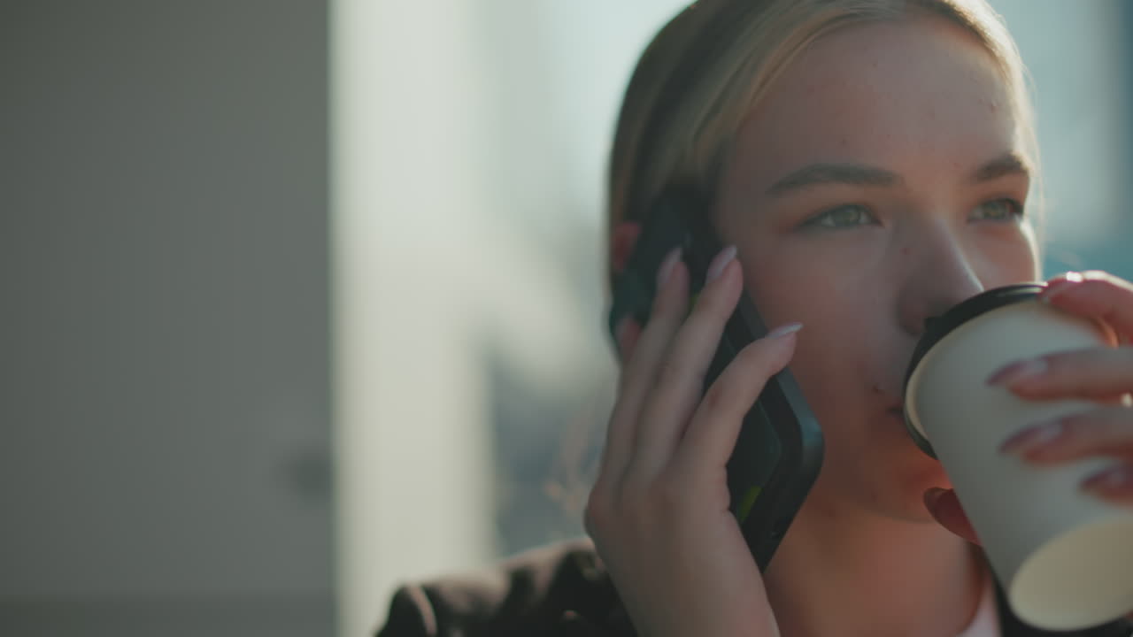 Close up of young lady on phone call walking as sunlight reflects off her hair and blur background, sipping coffee with calm expression, dressed in professional attire under bright daylight