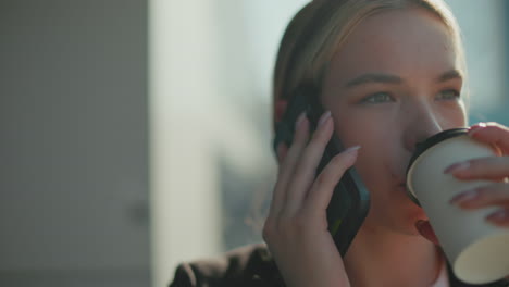 close up of young lady on phone call walking as sunlight reflects off her hair and blur background, sipping coffee with calm expression, dressed in professional attire under bright daylight
