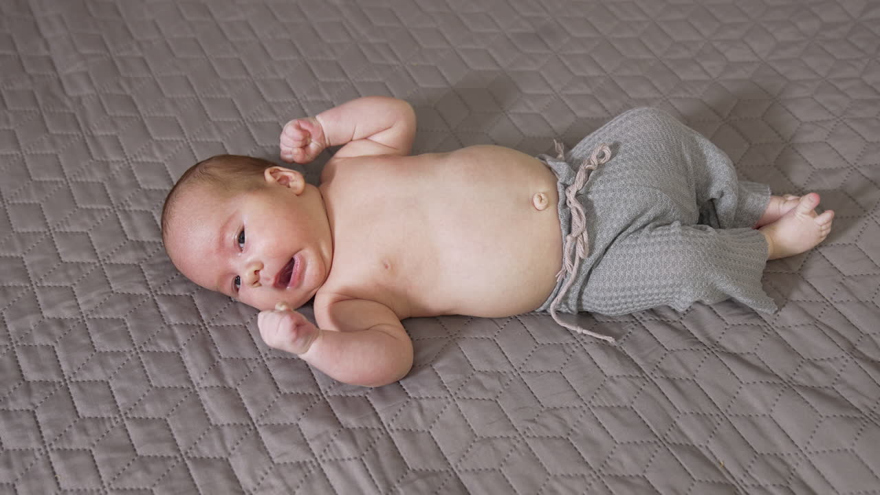 Little boy wearing grey pants lies on the bed. Cute newborn sneezes funnily. View from above on the child with naked belly.