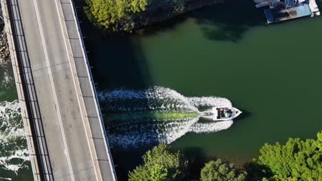 aerial drone footage shows a speedboat passing beneath a road bridge on a sunny day, surrounded by greenery and waterfront homes in gold coast