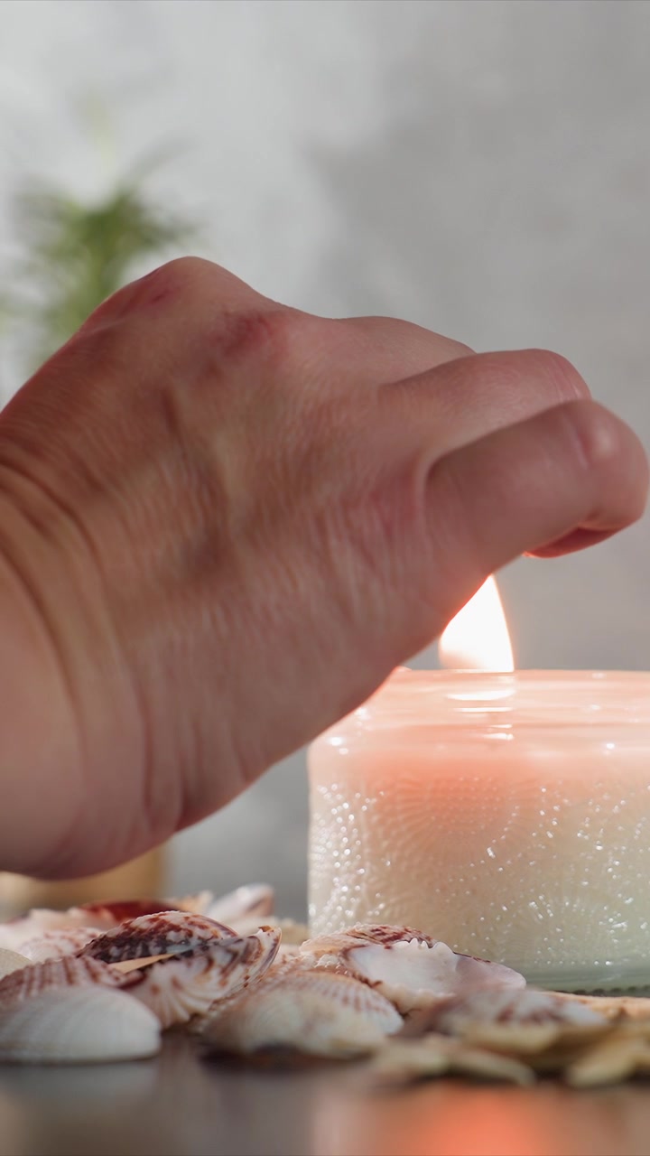 Primo piano di una mano che accende una candela inodore circondata da conchiglie su una superficie liscia con luce naturale soffusa. Il vaso di vetro, la cera testurizzata e le conchiglie decorative creano una calma scena costiera con un'atmosfera calda.