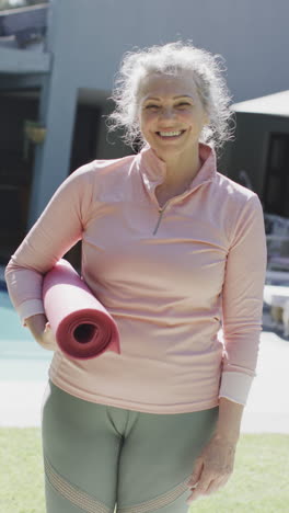 vertical video portrait of senior caucasian woman holding yoga mat in garden, slow motion