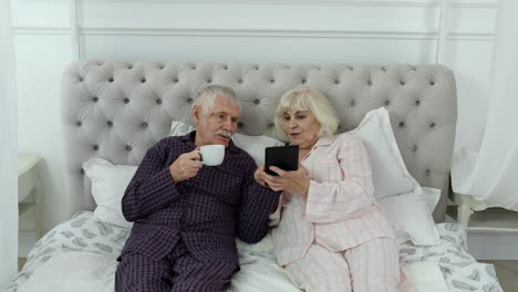 Senior-elderly-couple-wearing-pyjamas-lying-on-bed-looking-on-digital-tablet-laughing-and-having-fun