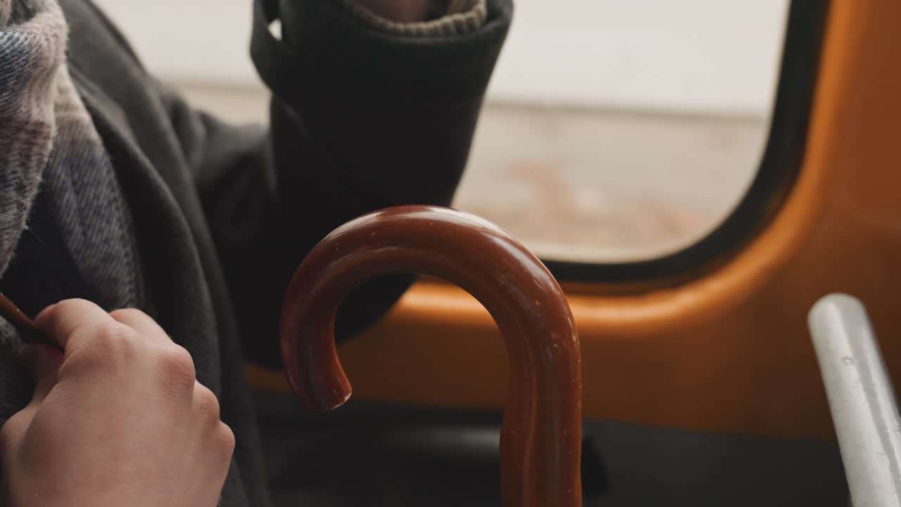 Mani su un bastone di legno, finestrino dell'autobus, un creatore che riflette durante un lento tragitto, primo piano di sciarpa e cappotto, luce naturale calda, impugnatura del bastone usurata, leggero movimento della mano, tranquilla vista urbana all'esterno.