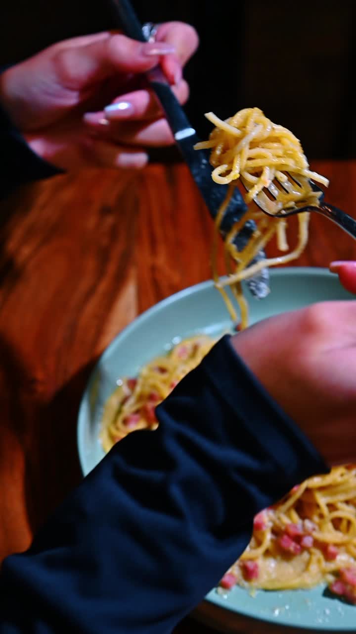 Primo piano di una persona che mangia spaghetti alla carbonara con forchetta e coltello. Mano con anelli che tiene le posate per mangiare pasta italiana con pancetta da una ciotola blu su legno.