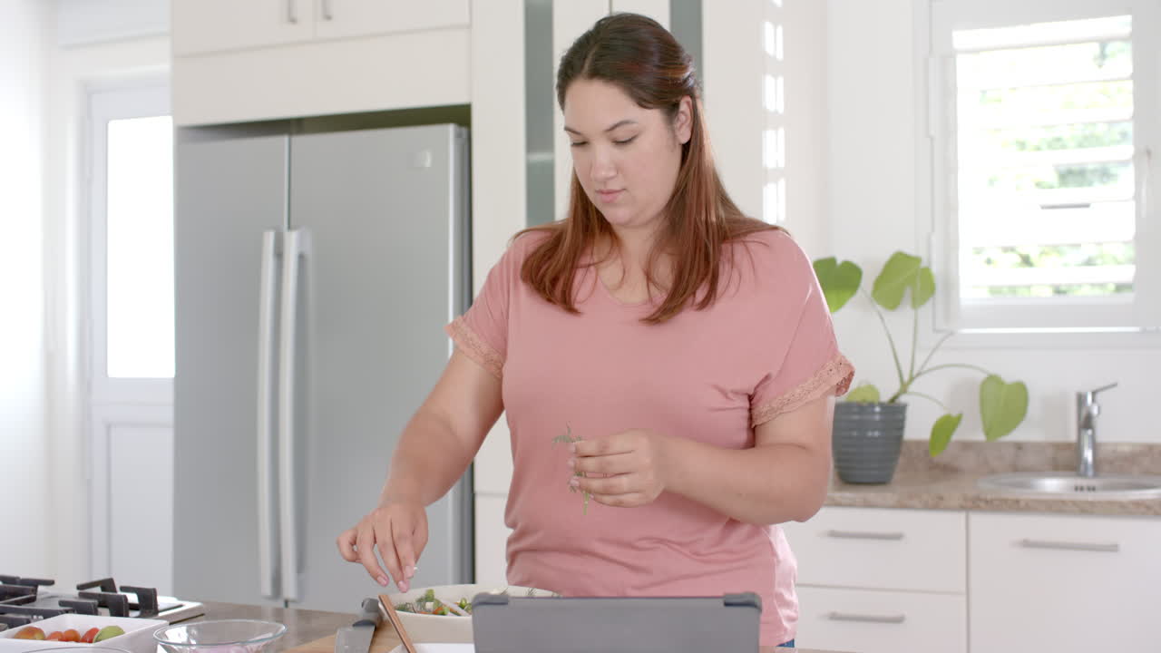 Felice donna birazziale di taglia forte che prepara un'insalata di verdure in cucina e usa un tablet, al rallentatore