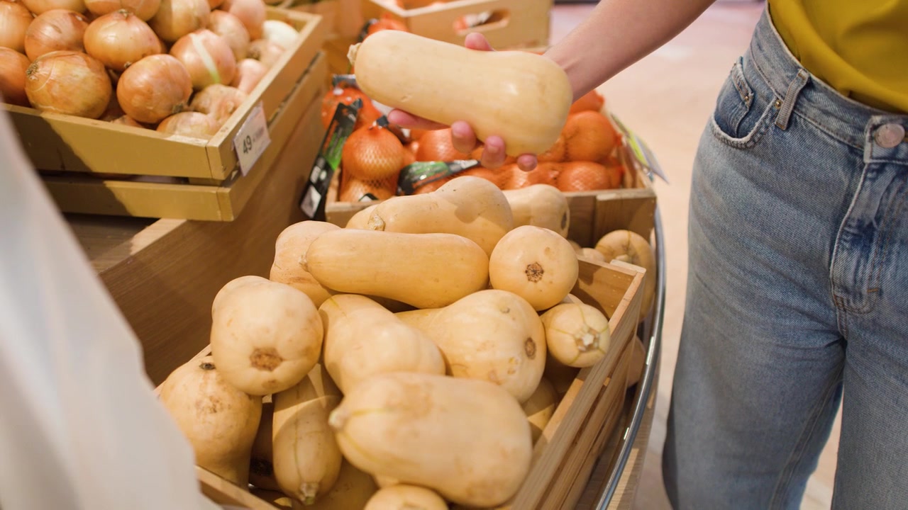 A shopper inspects and selects fresh butternut squash from a wooden bin in a supermarket