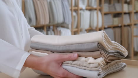 close-up of hands holding a stack of neatly folded neutral-colored textiles in a store or organized space