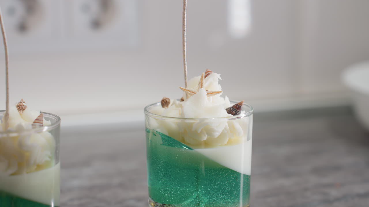 Professional baker places edible shell and sprinkle decorations on iced cake inside transparent dessert cup, adding final artistic touch with precision and elegance in bright modern kitchen setting