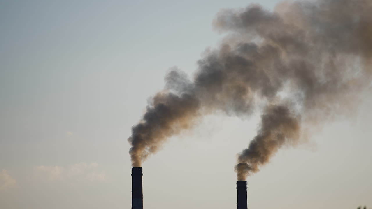 Tops of two plant chimneys polluting air with dark grey smoke. Environment pollution concept. Grey clear sky backdrop.
