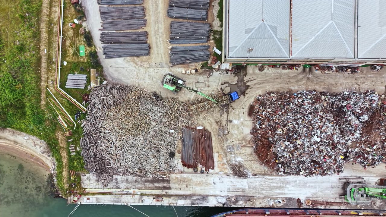 Vista di un ampio piazzale con cumuli di rottami metallici e materiali in legno, vicino a uno specchio d'acqua, durante le ore diurne in una trafficata zona industriale.