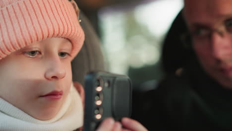 a close-up shot of a young girl wearing a pink beanie and jacket, sitting in a car and holding a phone, while a man, likely her father, leans in closely with a focused expression