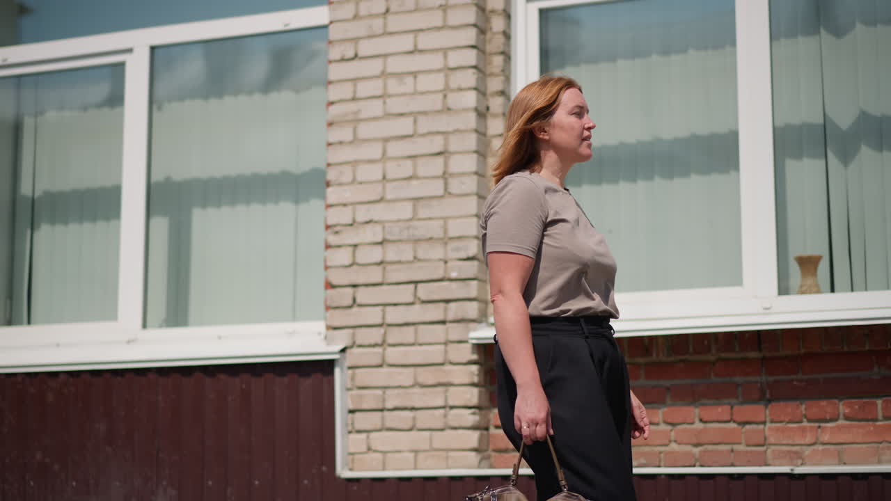 Side view of lecturer walking out of building holding handbag, hair gently swaying in wind under bright sunlight, face calm and thoughtful, representing peaceful professional moment