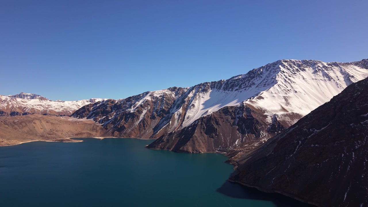 Volo planato lungo un profondo bacino idrico turchese, delimitato da aspre creste andine innevate e ripidi pendii rocciosi, sotto un cielo invernale limpido e blu. Embalse del Yeso, Cile.