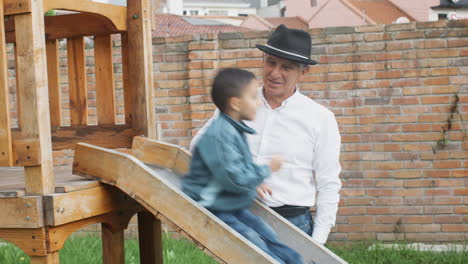 a tender moment between grandfather and grandson as they play together in a wooden playhouse with a slide in the backyard.