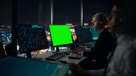 female air traffic controller with headset works on computer with green screen mock up in airport tower. office room with displays with navigation screens, airplane flight radar data for the team.