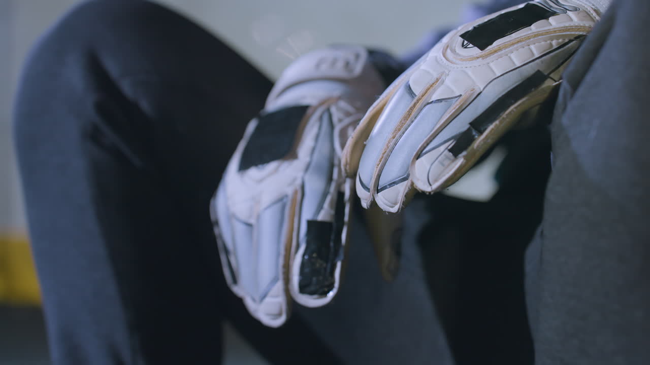 Close-up of hands wearing white sports gloves resting on knees of person sitting in casual sportswear, yellow marking on metallic fence in background