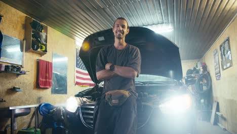 Portrait-of-a-confident-motorist-with-collected-hair-in-a-gray-T-shirt-who-looks-at-the-camera-and-leans-on-the-hood-of-his-car-in-his-garage-workshop