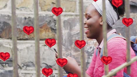 animation of red heart balloons over happy african american woman using smartphone in city