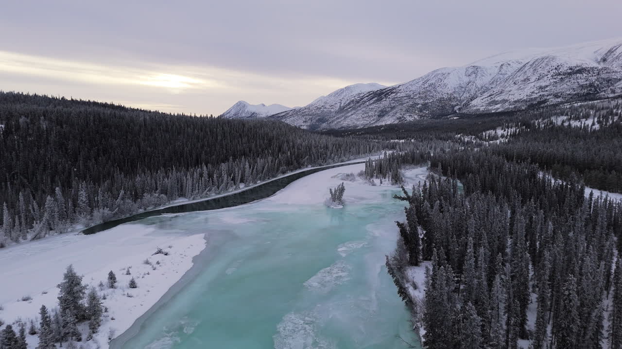 A wide-angle dolly forward shot flies over the Takhini River and snow-laden trees at sunset with majestic mountains in the background.