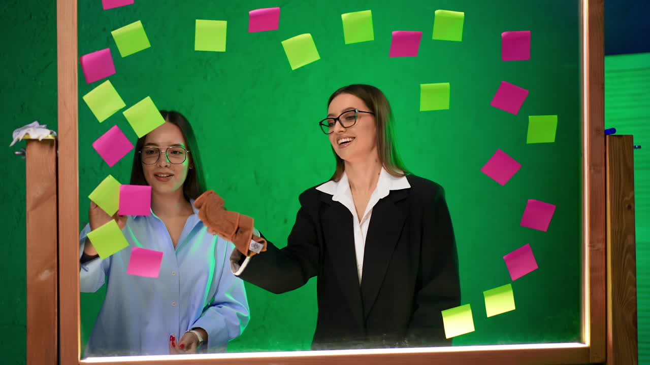 Positive ladies stand behind the glass board. Woman in black jacket cleans the board among the stickers.