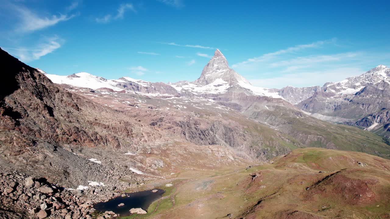 4K-Drohnen-Luftaufnahme des ikonischen Matterhorns, das sich über felsigem Alpgelände und fernen schneebedeckten Gipfeln erhebt und die dramatische Berglandschaft der Schweiz unter klarem blauem Himmel präsentiert.